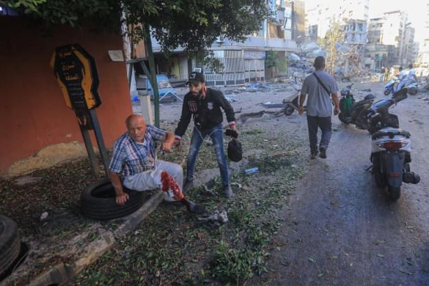 An older man sits on a sit walk. His lower leg is bleeding and a younger man holds his arm.
